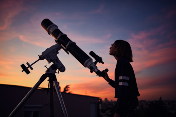 Person standing beside a telescope looking at a sky at sunset.