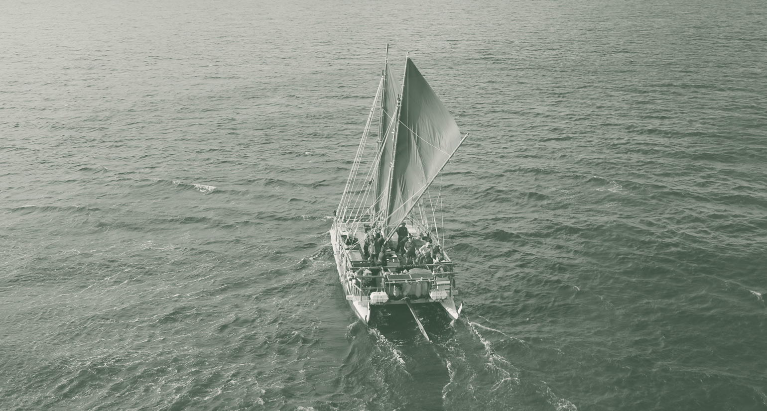  A double hulled waka is pictured from a birds-eye view above a calm sea. People can be seen on the deck below two large sails