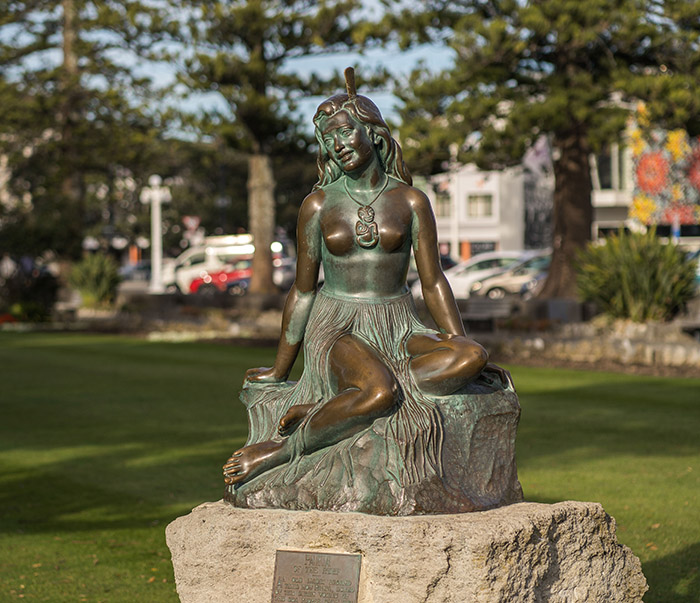 Bronze statue of Pania - a wāhine māori wearing a heru (bone comb), tiki pounamu necklace, and a grass piupiu (skirt), sitting on rock.