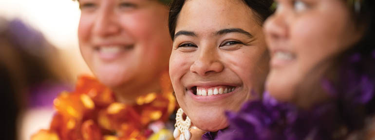 Decorative image: 3 people - the ones on either side wear lei and flower crowns, while the person in teh centre smiles at the camera. 