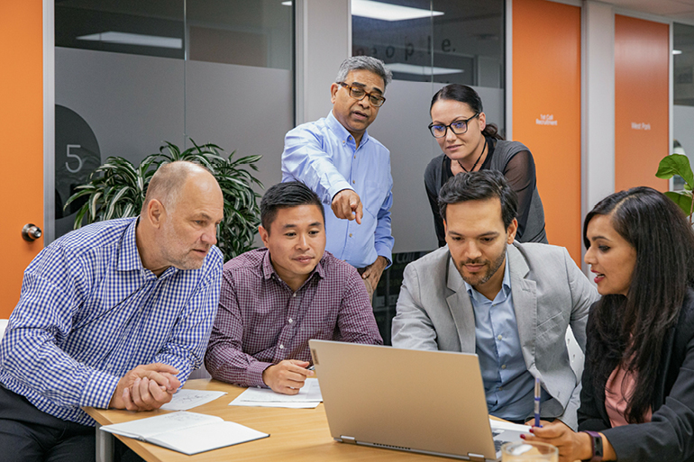 Four people seated round a table looking at a laptop while two people stand behind