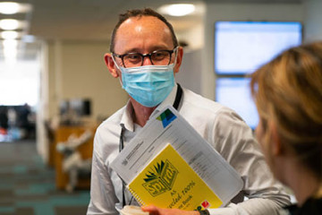 Photo of a man with glasses and wearing a mask, in an office environment, carrying papers and a notebook in conversation with someone on his left (they are only partially visible).