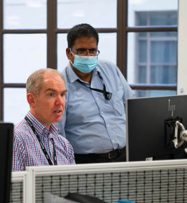 One man sitting at desk looking at a computer, another man with a mask stands next to him, looking over his shoulder at the computer screen.
