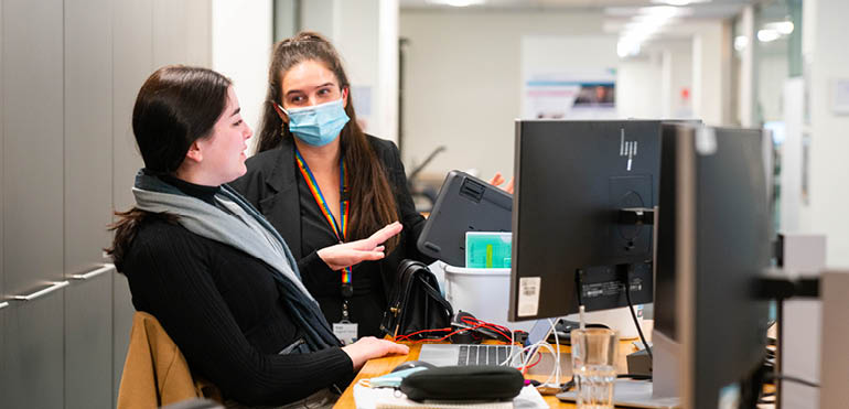 2 young women at a desk. One is sitting at the desk and talking she is not wearing a mask. The second young woman is standing listening to her. She wears a mask.