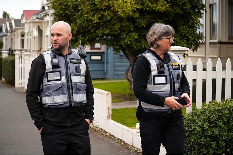 2 people walking in a suburban street, both wear uniforms of tenancy compliance officers. The man is looking out of frame to his right, the woman is talking and looking at something on her left.