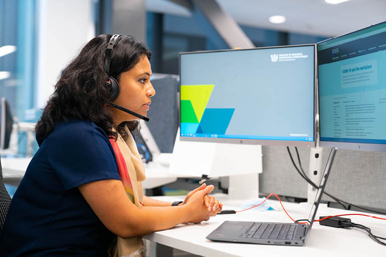 Young woman wearing a headset, taking a call. She is sitting at a desk with computer monitors in front of her.