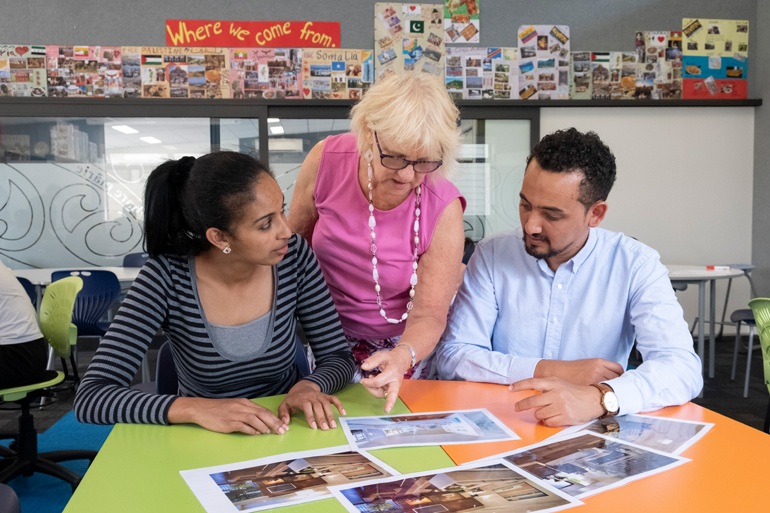 Two people sitting and being being shown some information by another person.