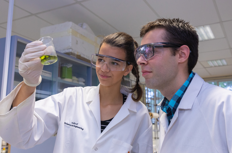 Two people in lab coats and safety glasses looking at a solution in a beaker.