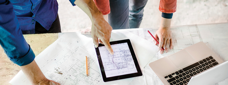 Decorative image: the hands of two people, one leaning on a printed architectural floorplan and the other pointing at a table showing another architectural floorplan