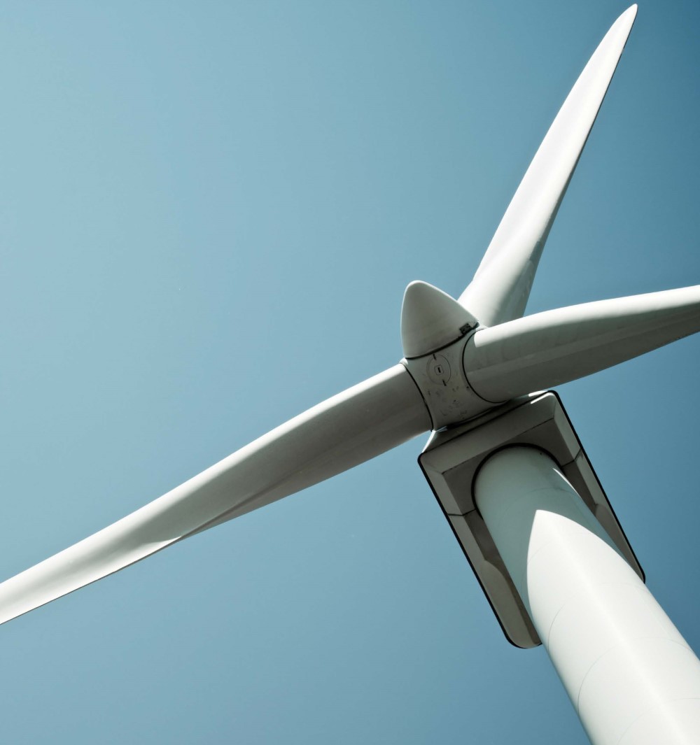 Photo of a wind turbine. The photo is taken from below near the base of the tower, showing the blades of the wind turbine against a clear blue sky.