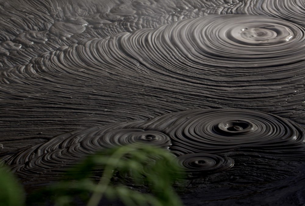 Photo of a mud pool. We see a number of concentric circles in the mud and some steam, showing it has been bubbling from steam coming from under the ground.