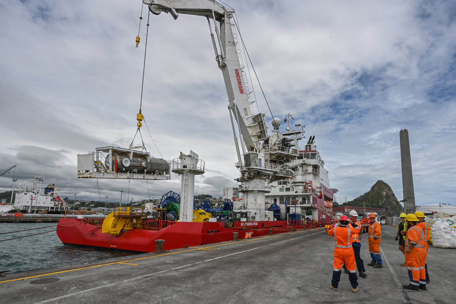 The Southern Star at the Port of Taranaki. 6 people in helmets and orange safety gear stand in the foreground while a hyperbaric reception facility being offloaded via crane in the background.