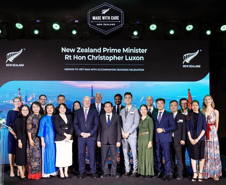 Prime Minister Rt Hon Christopher Luxon stands with 19 other people on a stage in front of a large video display. The display has the words "Made with care - New Zealand", the New Zealand fern mark, the prime minister's title, and "Mission to Viet Nam with accompanying business delegation".