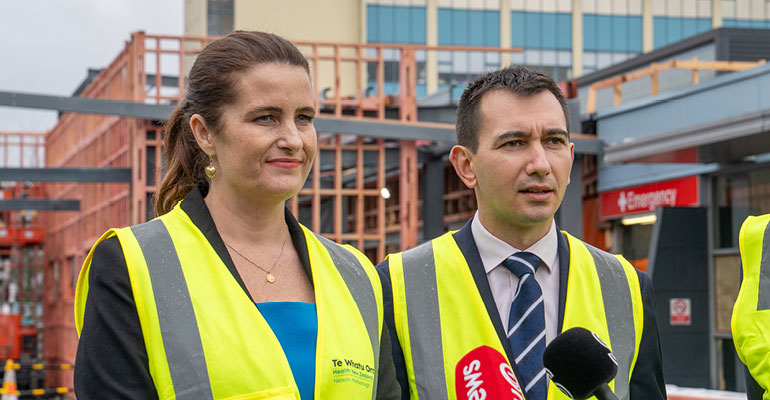 Ministers Nicola Willis and Simeon Brown being interviewed outside Nelson Hospital. The background shows building construction adjacent to the existing emergency department, and a taller building in the background.