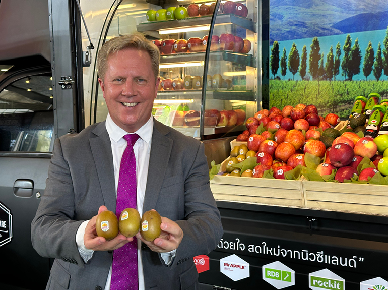 Minister for Trade and Investment, Hon Todd McClay, smiling at the camera and holding 3 kiwifruit. Todd is standing in front of a display of fruit from New Zealand featuring apples, kiwifruit, avocado, and possibly more. The display is mounted on the side of a vehicle which is obscured.