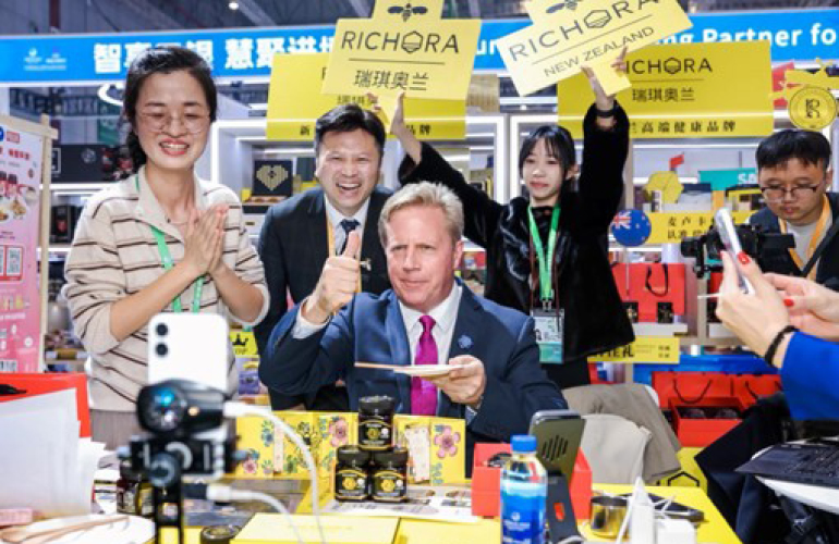 Hon Todd McClay in a brightly lit shopping mall type of space, giving a thumbs up to a mobile phone camera and holding a plate. Todd is surrounded by four other people clapping, laughing, and holding signs relating to the product being promoted.