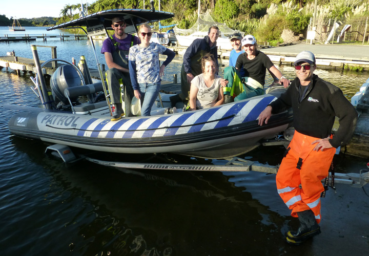 Photo: A patrol dinghy with 6 people in it, sitting on the edge of the water. One person in the foreground is leaning on the boat and smiling. He is wearing orange pants and a blak shirt. 