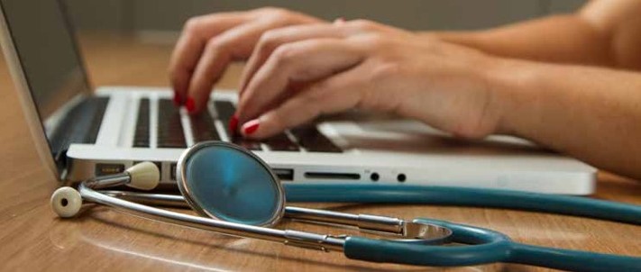 Cropped image of person's hands typing on laptop with stethascope sitting next to laptop.