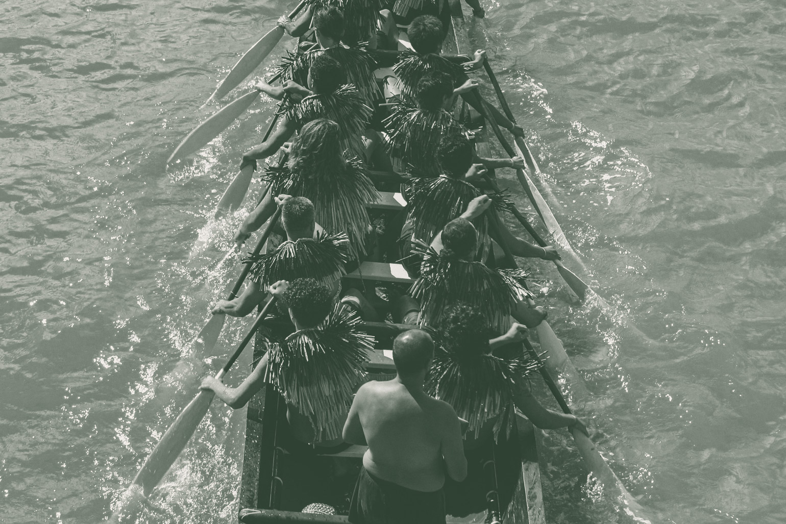 A birdseye view of 10 Kaihoe waka paddlers comprising men and women in traditional Maori dress propelling a two person wide waka forward in the water.