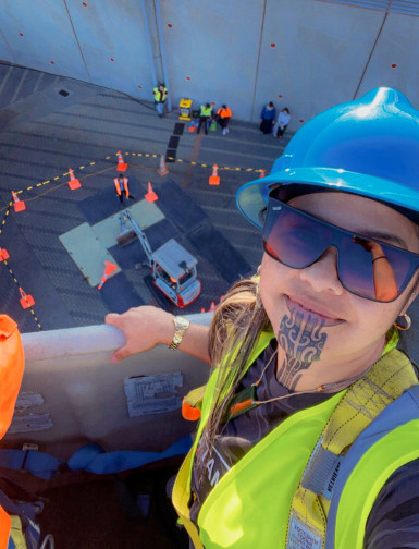 Wahine with moko kauae wearing hard hat and hi-vis vest on a cherry picker. 