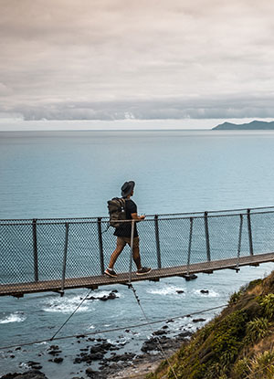 A person walks over a swing bridge on the Kapiti Escarpment walkway with Kāpiti Island in the background