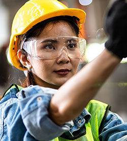 A young woman in hard hat, goggles and high vis vest working in a factory.