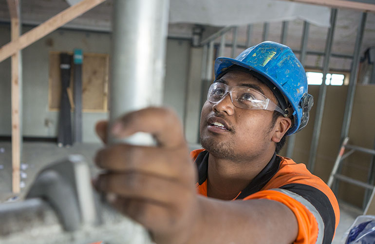 Man wearing an orange hi-vis vest and blue hard hat holding on to some scaffolding