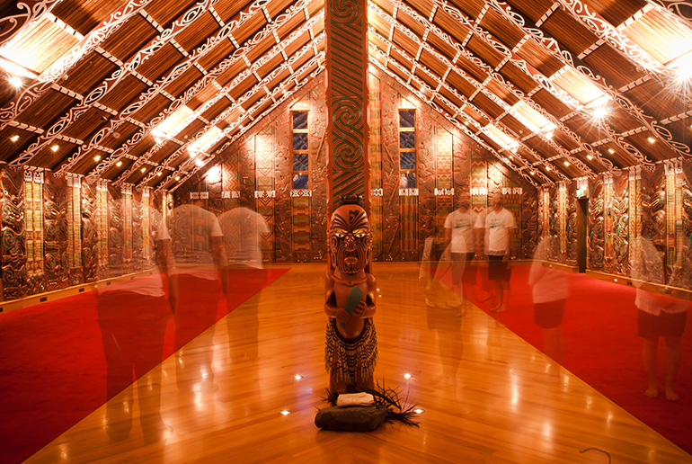 Inside view of a marae looking directly at the center pou carving with blurred images of people gathering inside