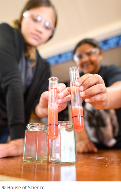 Two young rangatahi women in safety goggles compare test tubes © House of Science