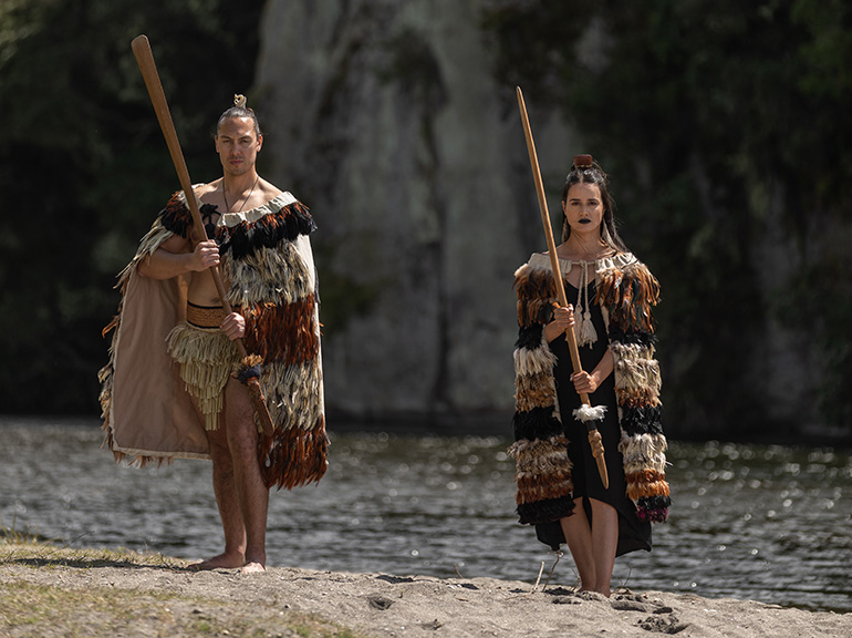 A man and woman in traditional Māori clothing and holding taiha stand on the shores of Lake Taupō