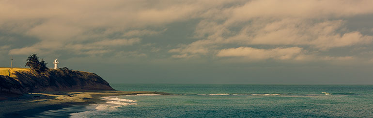 Sun strikes a small white lighthouse on a peninsula as the sea breaks below