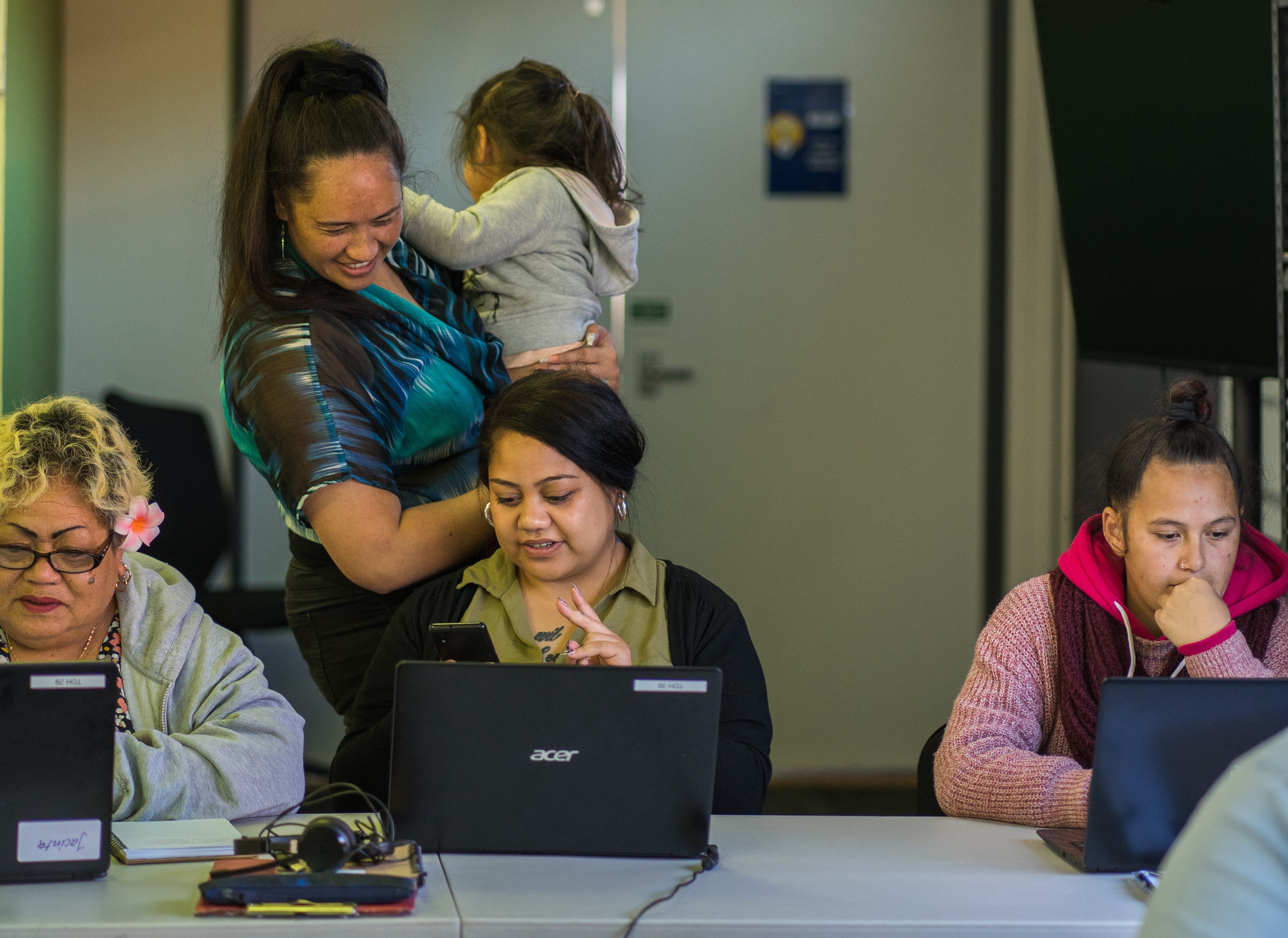 A wāhine standing holding her toddler looks down at another wāhine sitting and working on her laptop