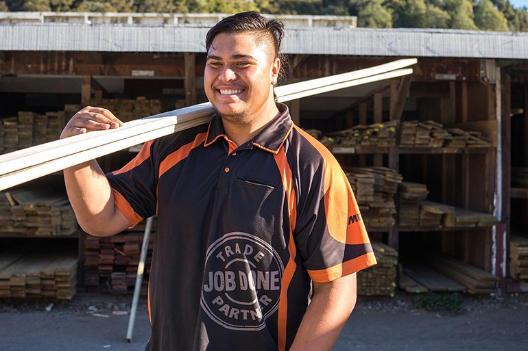 Young male smiling, carrying two pieces of timber in a timber yard.