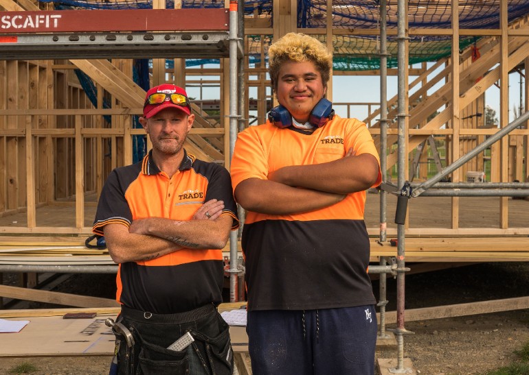Young man and man standing, folding their arms with a house being built behind them.