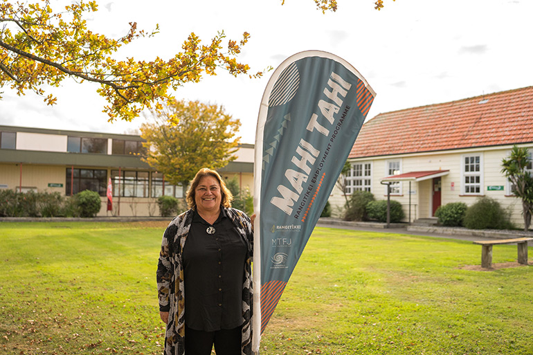 A woman standing by a sign that reads ‘Mahi Tahi’ with buildings in background.