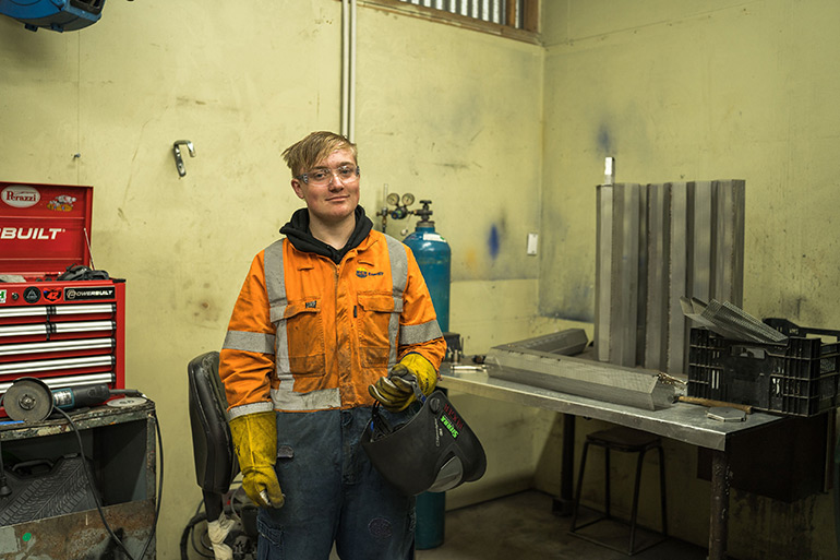 Young man wearing safety overalls and gloves carrying a welders helmet.