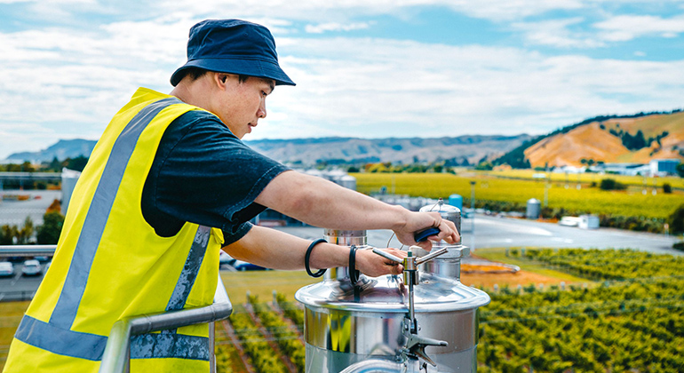 A man in a high vis vest adjusts an outdoor wine tank.