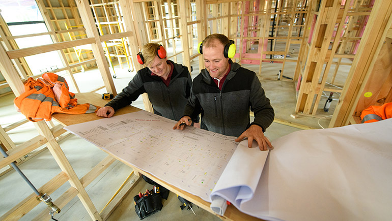 Two people looking at building plans on a table inside a house under construction.