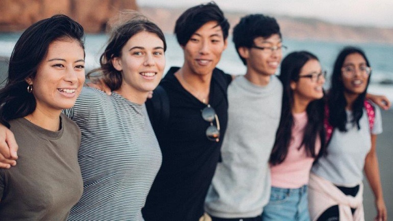 Six young people of different ethnicities and gender in a line hands over each others shoulders with a beach in background.