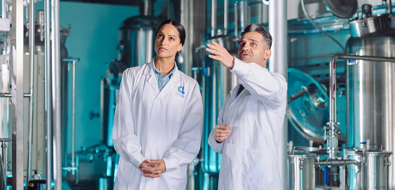 Man and Women in a factory in work coats with man gesturing to something.