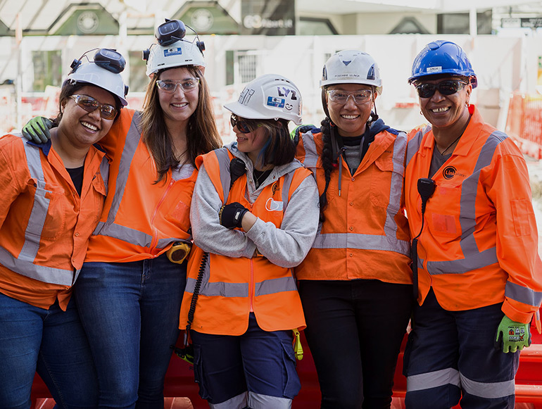 Women standing together smiling. They are wearing hard hats, safety glasses, and hi vis vests.