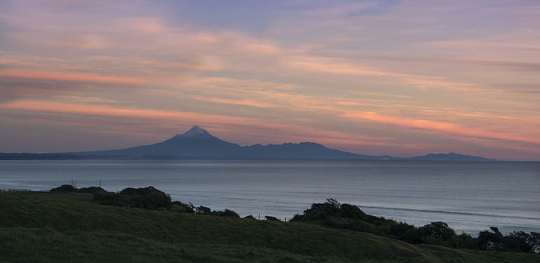 Sunset over mount taranaki taken from a distance, with ocean and land in the foreground.