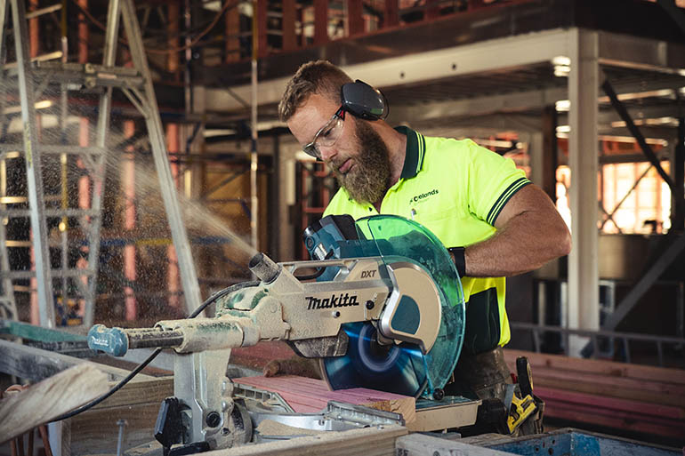 A male builder using a skillsaw wearing earmuffs, eye protection and a high viz shirt. 