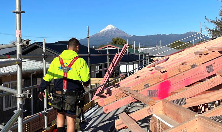 Construction worker strapped to a harness on a scaffolding, on top of a roof frame, with mount taranaki in the background
