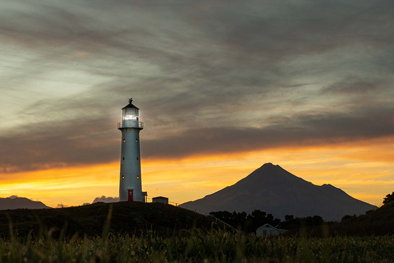 Cape Egmont Lighthouse with Mount Taranaki and a sunrise behind it.