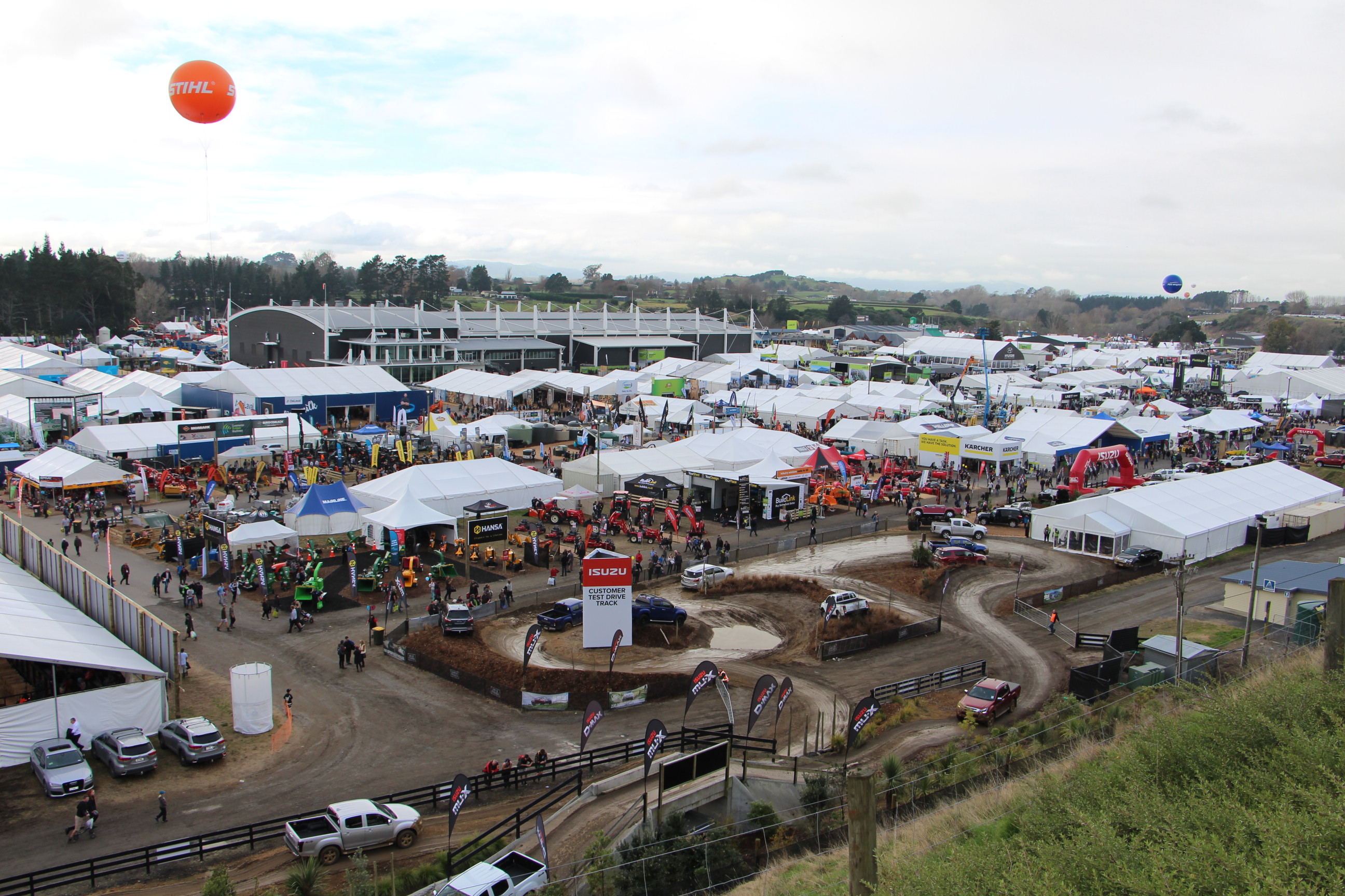 Aerial view of many tents, people and farm machinery. In the foreground are several four wheel drive utes driving along a muddy track