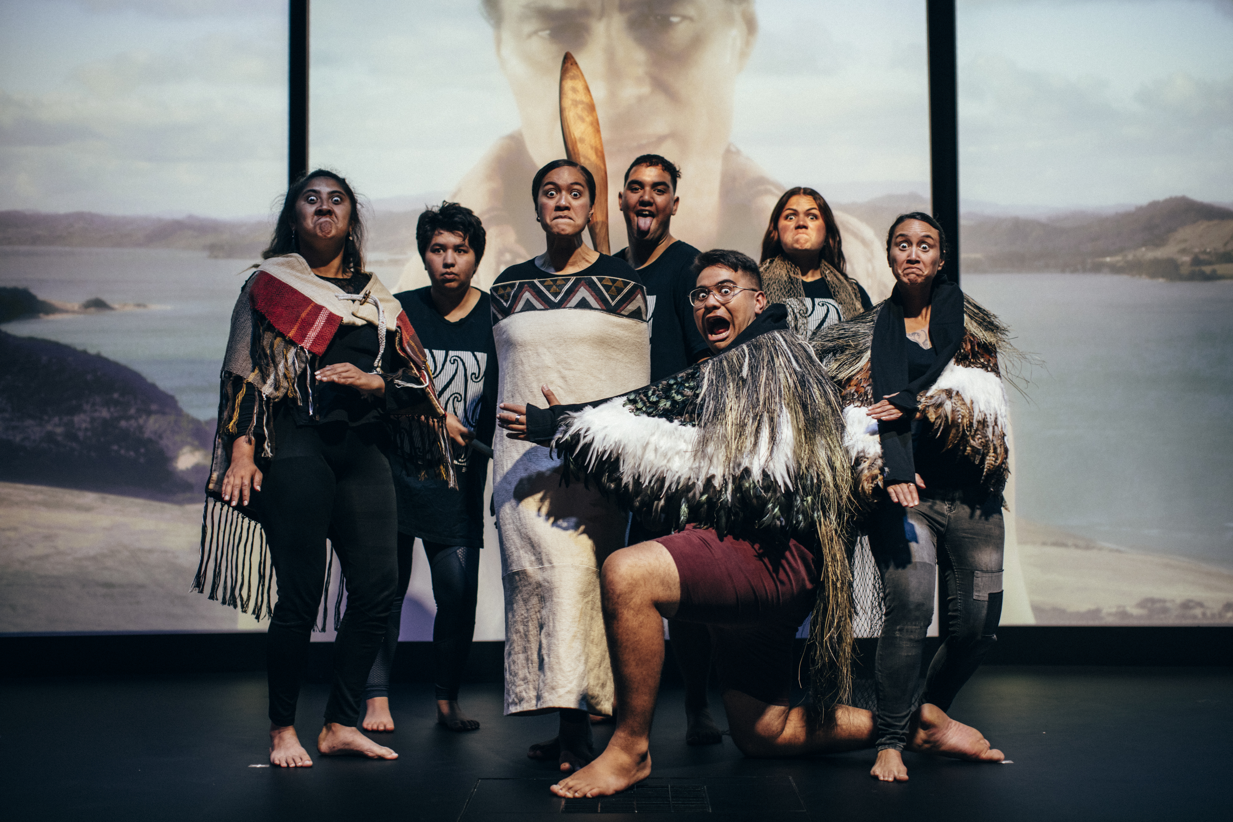 Several Māori youth grouped closely together. All are wearing a mix of modern and traditional clothing, and are performing a pūkana