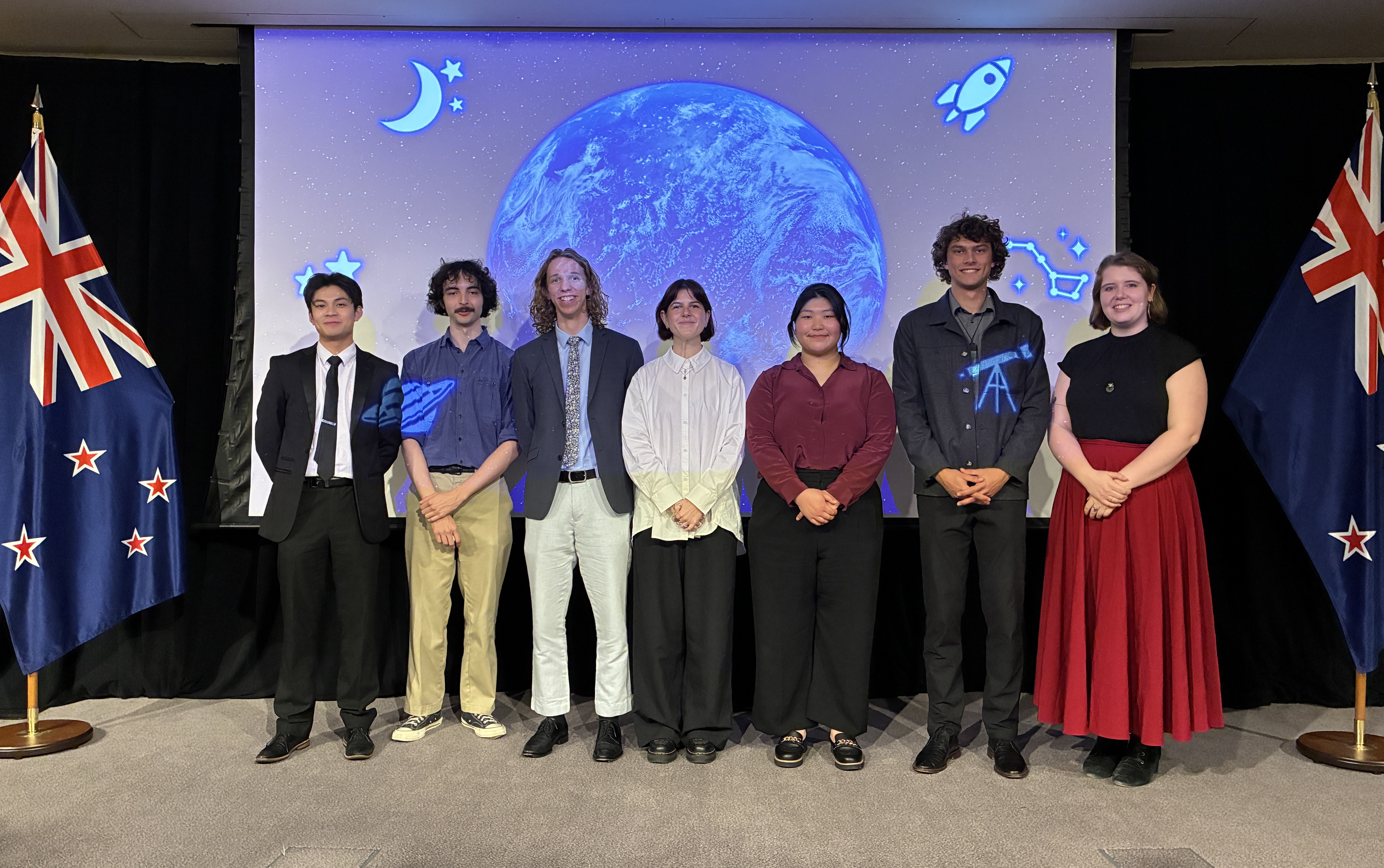 2026 New Zealand Space Scholarship recipients standing on a stage in front of of a screen with an image of the world