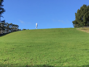 A mobile tower on top of a hill surrounded by green grass, trees and blue sky.