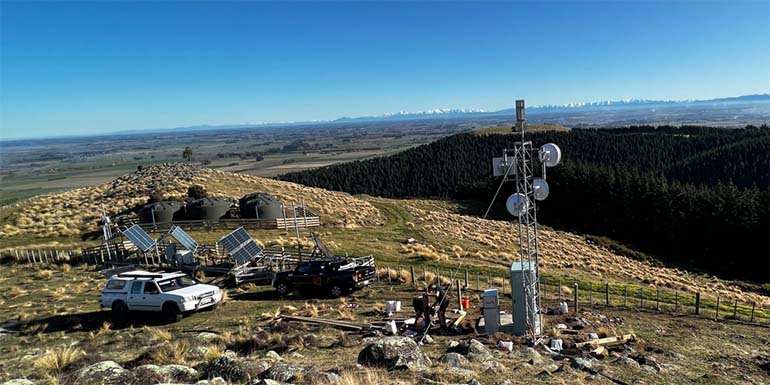 White cars parked on crest of hill, solar panels and a mobile tower surrounded by rocky terrain and grass